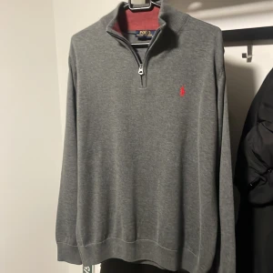 Grå tröja med dragkedja från Polo Ralph Lauren - Säljer en stilren grå tröja från Polo Ralph Lauren med en halv dragkedja och röd logga på bröstet. Perfekt för både vardag och lite finare tillfällen. Tröjan har en bekväm passform och är i mycket bra skick. Passar perfekt till höst och vår!