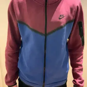 Säljer en snygg blå och vinröd Nike tech  hoodie i storlek S. Hoodien har en dragkedja framtill och en praktisk ficka med dragkedja på ärmen. Perfekt för höst och vår med sin sportiga stil och bekväma passform.