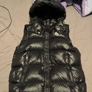 Svart dunväst från Moncler - Säljer en snygg svart dunväst från Moncler i mycket bra skick. Västen har en quiltad design med dragkedja och en praktisk huva. Perfekt för höst och vinter för att hålla värmen med stil. Passar både till vardags och lite finare tillfällen.
