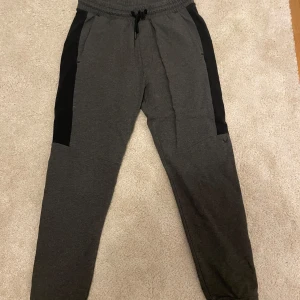 Grå mjukisbyxor från SOC - Säljer ett par grå mjukisbyxor från SOC i storlek S. Byxorna har svarta sidopaneler och en bekväm resårmidja med snörning. Perfekta för en avslappnad dag hemma eller lätt träning. De är i bra skick och redo för nya äventyr!