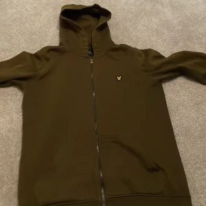 Grön hoodie från Lyle & Scott - Säljer en snygg grön hoodie från Lyle & Scott i storlek 12/13 år. Den har en dragkedja framtill och en liten logga på bröstet. Perfekt för höst och vår, med långa ärmar och en skön huva. Passar både till skolan och fritiden!