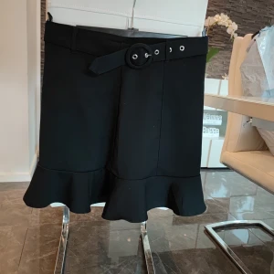 Svart kjol med volang och bälte - Snygg svart kjol med volangdetalj längst ner och ett matchande bälte i midjan. Perfekt för både vardag och festliga tillfällen. Kjolen har en stilren design och är lätt att matcha med olika toppar.