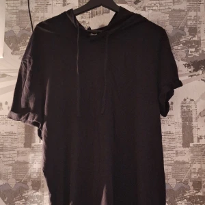 Svart T-shirt med luva - Säljer en svart t-shirt med luva i bra skick. Den har korta ärmar och en klassisk huva med snörning. Väl använd och relativt sliten men tyget i sig var redan slitet från början.