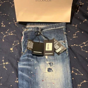Blå jeans från Dsquared2 - Säljer ett par snygga blå jeans från Dsquared2 i modellen Skater Jean. De har coola slitningar och färgstänk som ger en unik look. Perfekta för en avslappnad stil med en touch av edge. Helt nya med tags kvar! 100% äkthet från THERNLUNDS Stockholm köpta i Mal of scandinavia i Solna.