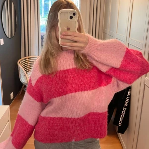Rosa och röd randig stickad tröja - Supermysig stickad tröja i rosa och rött med breda ränder. Perfekt för kyliga dagar när du vill vara både varm och stilren. Tröjan har en avslappnad passform och långa ärmar, vilket gör den idealisk för både höst och vinter och sommar. Ett måste i garderoben för den som älskar färg!