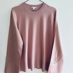 Rosa tröja med skjortärmar från COS - Säljer en stilren rosa tröja från COS med långa skjortärmar och knappar vid ärmsluten. Tröjan har en rund halsringning och är tillverkad i ett mjukt material som ger en bekväm passform. Perfekt för en avslappnad look. Nyskick, endast använd en gång