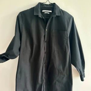 Fifth avenue lång skjorta med knytband - Svart skjorta/blus med långa ärmar, vidd och knytband i ryggen. Dekorativt band på ärmarna. Skjortan har en klassisk krage och knappar framtill. Bröstficka på ena sidan. Längd till ovanför knäna. Slitsar i sidorna. 96% bomull, 4% elastan. Tvätt 40 grader. Perfekt för en stilren look.