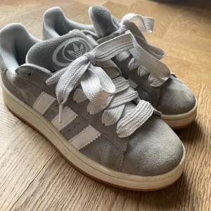 Grå Adidas Campus sneakers - Snygga grå Adidas Campus sneakers i mocka med vita detaljer och breda snören. Perfekta för en stilren look med den klassiska tre-randiga designen på sidan. Skorna har en bekväm sula och är ett måste för alla sneaker-älskare.