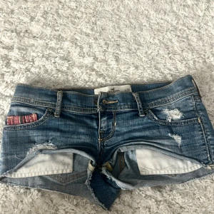 Blå jeansshorts från Hollister - Snygga blå jeansshorts från Hollister med slitna detaljer och låg midja. Perfekta för en avslappnad sommarlook. Shortsen har klassiska fem fickor och knappgylf. (Förra ägaren bilder❤️)