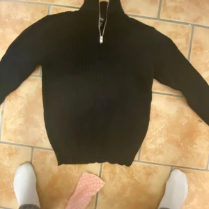 Svart ribbad tröja med dragkedja - Säljer en svart ribbad tröja med dragkedja vid halsen. Tröjan har långa ärmar och en hög krage, perfekt för kyligare dagar. Enkel och stilren design som passar till det mesta.