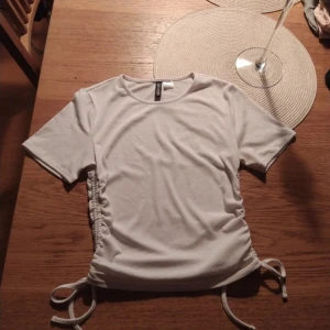 Ljus Beige kortärmad topp från H&M Divided - Säljer en ljus beige kortärmad topp från H&M Divided. Toppen har snörning på sidorna som ger en snygg detalj och gör den justerbar i passformen. Perfekt för en stilren look.