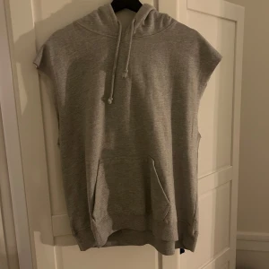 Grå ärmlös hoodie - Säljer en grå ärmlös hoodie med luva och dragsko. Perfekt för en avslappnad stil. Den har en stor ficka framtill och är gjord i mjukt material.