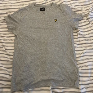 Grå t-shirt från Lyle & Scott - Säljer en stilren grå t-shirt från Lyle & Scott med ett broderat gult emblem på bröstet. Perfekt för en avslappnad look. T-shirten är kortärmad och gjord i mjukt bomullsmaterial.     14 till 15 åringar