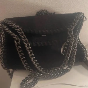 Svart axelväska med kedjedetaljer - Snygg svart axelväska med coola kedjedetaljer runt kanterna och som axelrem. Väskan har en praktisk magnetknapp för enkel stängning och en ljusrosa insida. Perfekt för att ge din outfit en edgy touch. Kommer inte gå ner i pros