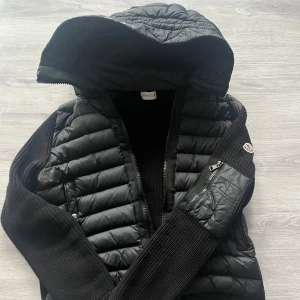 Svart quiltad jacka från Moncler - Snygg svart quiltad jacka från Moncler med dragkedja och stickade ärmar. Jackan har en hög krage och en praktisk ficka med dragkedja på ärmen. Perfekt för kyligare dagar och ger en stilren look. Tejpad på luvan deför är priset billigare priset kan diskuteras fraktar inom 12 timmar 📦✅🚚