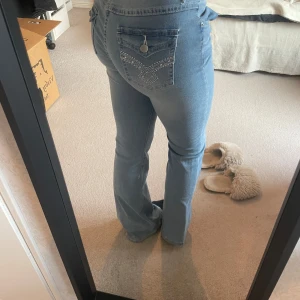 Blå bootcut jeans - Snygga blå bootcut jeans med klassisk femficksdesign och knappgylf. Jeansen har en lätt slitning vid midjan och dekorativa sömmar på bakfickorna.