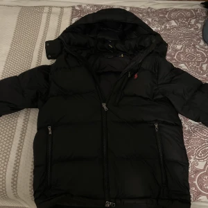  Polo Ralph Lauren jacka/ polo jacka - Säljer en svart dunjacka från Polo Ralph Lauren med huva och dragkedja. Jackan har långa ärmar och två framfickor med dragkedjor. Den ikoniska loggan syns på bröstet. Perfekt för kyliga dagar! Storlek M men passar även s