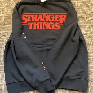 Svart Stranger Things tröja från Zara - Svart tröja från Zara med Stranger Things-tryck i rött på framsidan. Ärmarna har färgglada illustrationer av karaktärer från serien. Perfekt för fans av Stranger Things! 🖤Stl. 164.