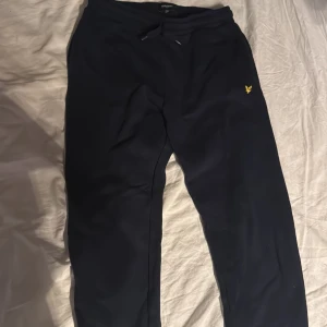 Marinblå mjukisbyxor från Lyle & Scott - Snygga svarta mjukisbyxor från Lyle & Scott med en gul logga på låret. Byxorna har en bekväm passform med snörning i midjan för justerbarhet. Perfekta för en avslappnad stil.