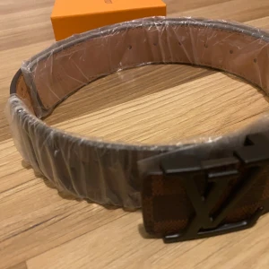 Brunt rutigt bälte från Louis Vuitton - Säljer ett stilrent brunt bälte från Louis Vuitton med ett klassiskt rutigt mönster. Bältet har en elegant LV-spänne i mörk metall. Perfekt för att ge en lyxig touch till din outfit. Kommer i originalförpackning.