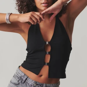 Svart halterneck topp med metallspännen - Helt ny, aldrig använd trendig svart halterneck topp från Gina med unika metallspännen framtill. Toppen är ärmlös och har en stilren design som passar perfekt för en modern look. Perfekt för att skapa en chic outfit.