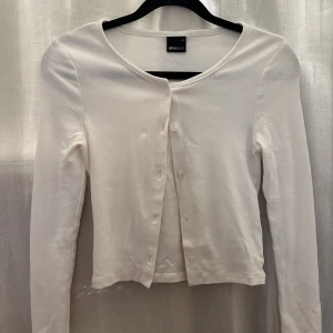Vit kofta från Gina Tricot - Säljer en stilren vit kofta från Gina Tricot i storlek XS. Koftan har långa ärmar och knappar framtill, perfekt för lager-på-lager-stil. Enkelt och klassiskt plagg som passar till många tillfällen.