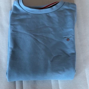 Ljusblå tröja från GANT - Säljer en stilren ljusblå tröja från GANT med rund halsringning. Tröjan har en liten broderad logotyp på bröstet. Perfekt för en avslappnad look.