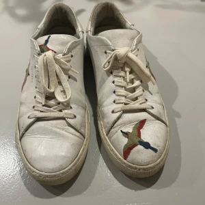 Vita sneakers med broderade fåglar från Axel Arigato - Snygga vita sneakers från Axel Arigato med färgglada broderade fåglar på sidorna och tån. Skorna har klassisk snörning och en stilren design som passar perfekt för en trendig look. Skick 7/10 pris är fast.