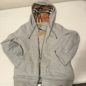 Grå hoodie från Burberry - Säljer en stilren grå hoodie från Burberry med dragkedja och klassiskt rutigt mönster i huvan. Perfekt för en avslappnad och trendig look. Hoodien har långa ärmar och en bekväm passform.