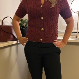 Vinröd kabelstickad kofta med korta ärmar - Säljer en vinröd kabelstickad kofta med korta ärmar och dekorativa guldknappar. Perfekt för en stilren look. Koftan har en klassisk design med ribbade kanter.