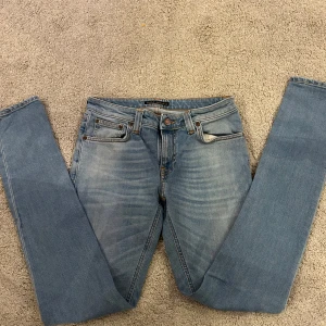 Blå jeans från Nudie Jeans - Snygga blå jeans från Nudie Jeans med klassisk femficksdesign och orange söm på bakfickorna. Jeansen har en slim passform och är tillverkade i denim. Perfekta för en stilren look.