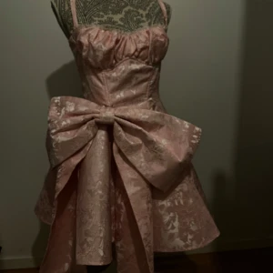 Rosa klänning med stor rosett - Charmig rosa klänning med blommigt mönster och en stor rosett i midjan. Klänningen har smala axelband och en kort längd som ger en söt och elegant look. Perfekt för speciella tillfällen. Rosetten är även justerbar vilket innebär att du kan ha den både fram och bak samt på andra klädesplagg oxå. Klänningen funkar även helt utan rosett. Den är helt oanvänd med prislapp kvar, säljer pga lite för stor storlek för mig