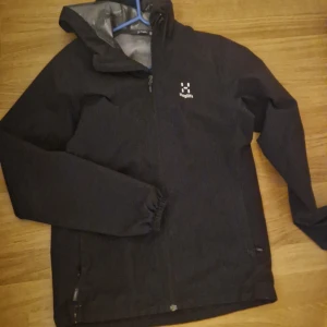 Svart vindjacka från Haglöfs - Säljer en svart vindjacka från Haglöfs med dragkedja och huva. Jackan har långa ärmar och är tillverkad i ett slitstarkt material. Perfekt för utomhusaktiviteter och skyddar mot vind och regn. Lite slitet där nere som ni ser på bilden men inget som syns annars är den i nytt skick 