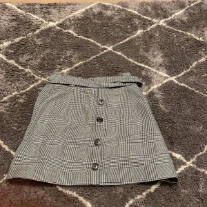 Rutig kjol med knappar - Snygg rutig kjol i svart och vit med knappar framtill. Kjolen har en klassisk design med ett bälte i midjan för extra stil. Perfekt för en chic look. Den har även två fickor som är väldigt praktiska.