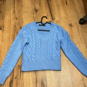 Blå stickad tröja med flätmönster - Säljer en snygg blå stickad tröja från Zara med ett klassiskt flätmönster. Tröjan har en v-ringad hals och långa ärmar, perfekt för kyligare dagar. Den är croppad och har ribbade kanter för en stilren look. Passar perfekt till jeans eller kjol!