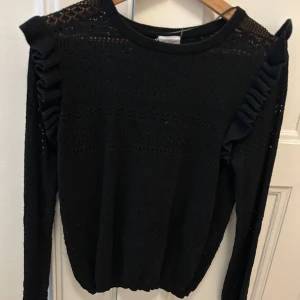 Svart tröja med volangdetaljer från Stories - Snygg svart tröja från Stories med volangdetaljer längs axlarna. Tröjan har ett fint mönster och är långärmad, perfekt för både höst och vinter. Den är i storlek S (36-38).