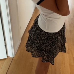 Blommig kortkjol - Söt kortkjol med blommigt mönster i svart och rosa. Kjolen har en volangdetalj som ger den extra charm och rörelse. Perfekt för en avslappnad och stilfull look.