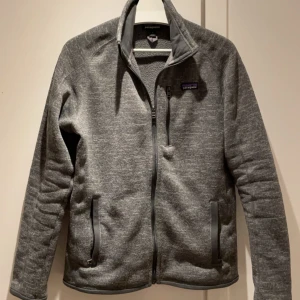 Grå Patagonia fleecetröja - Säljer en grå fleecetröja från Patagonia med dragkedja framtill och praktiska fickor. Jackan har en stilren design och är perfekt för kyligare dagar. En trendig tröja som är väldigt efterfrågad. Hör av er vid frågor!