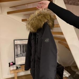 Svart parkajacka med pälsdetaljer från Hollies - Snygg svart jacka från Hollies med en fluffig pälsdetalj på huvan. Jackan har praktiska fickor och en dragkedja framtill. 