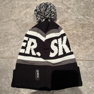 Svart och grå mössa från Planks - Snygg svart och grå mössa från Planks med texten 'SKIER' i vitt. Mössan har en stor tofs på toppen och är perfekt för kalla dagar. Den är ribbad och har en uppvikt kant för extra stil och värme.