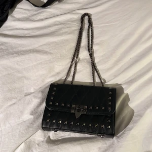 Svart axelväska med nitar - Snygg svart axelväska med kedjeaxelrem och coola nitar. Väskan har ett quiltat mönster och ett lås framtill. Perfekt för att ge din outfit en edgy touch.