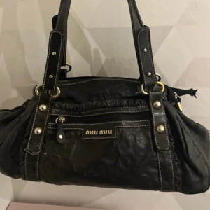 Svart skinnväska från Miu Miu - GÅR INTE NER I PRIS! Snygg svart skinnväska från Miu Miu med dubbla handtag och dragkedja. Väskan har en praktisk ytterficka med Miu Miu-logga och ett rutigt innerfoder. Snygg till alla outfits. Den har totalt 4 fack och på ett fack saknar den en dragkedja och sömmen inuit facket har gått upp vilket gör att saker kan ramla in i väskan, men det går lätt att sy själv och är ingenting som synd på utsidan, därför skulle jag säga att väska fortfarande är i god skick! 