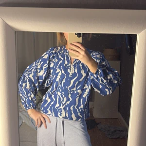 Blå och vit mönstrad blus - Säljer en blå och vit blus med ett vågigt mönster. Blusen har långa ärmar och en lös passform, perfekt för en avslappnad stil. Den har en v-ringad hals och är tillverkad i ett lätt material.