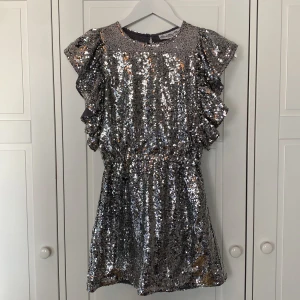 Glittrig silverklänning från How to Kiss a Frog - Säljer en superfin silverglittrig klänning från How to Kiss a Frog. Den har volangdetaljer på ärmarna och en söt passform med något intagen resårmidja. Perfekt för festliga tillfällen när du vill glänsa lite extra! På några ställen har paljetterna lossnat, därav nedsatt pris.