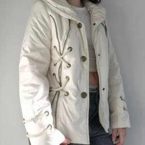 Vit jacka med snörning från Rosse Line - Säljer en snygg crèmefärgad jacka från Fornarina i mycket bra skick. Jackan har coola snörningsdetaljer och knappar framtill. Kragen kan vikas upp. Den är långärmad och har en normal passform. Perfekt för höst och vår med sin stilrena design!