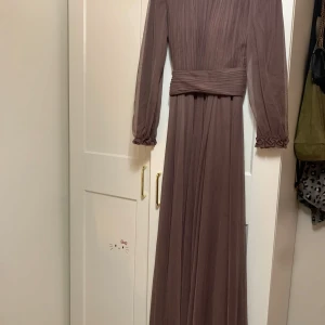 Smutslila långklänning från Mihir - Elegant brun långklänning från Mihir med plisserad design och långa ärmar. Klänningen har en hög krage med fjäderdetaljer och ett brett midjeband som ger en smickrande passform. Perfekt för höst- och vinterfester!