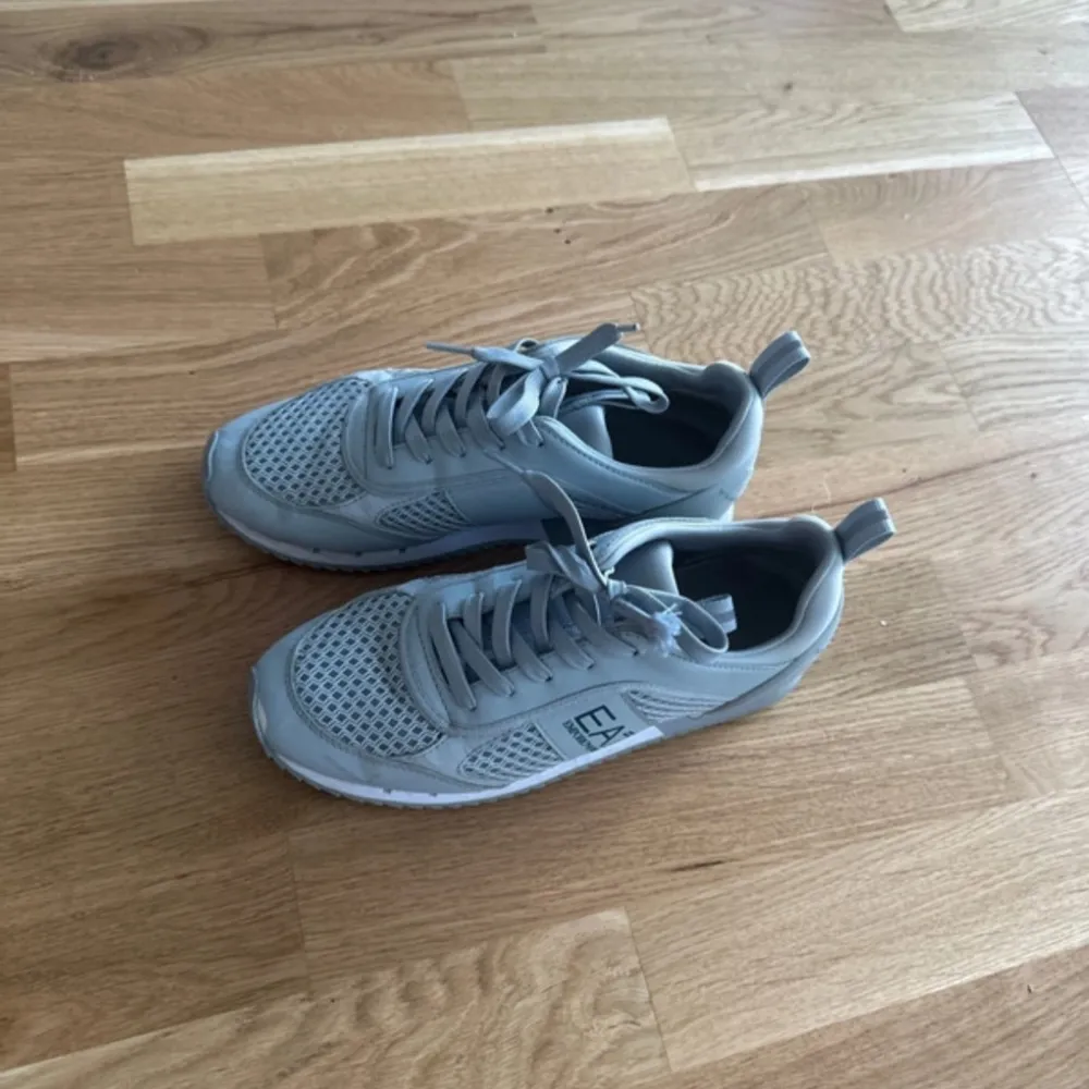 Säljer ett par snygga grå sneakers från EA7 i mycket bra skick. De har en stilren design med snörning och är perfekta för både vardag och träning. Skorna är gjorda i syntetmaterial med en luftig mesh-överdel som ger bra andningsförmåga. Perfekta för vår och sommar!. Skor.