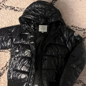 Svart glansig dunjacka från Moncler - Säljer en snygg svart glansig dunjacka från Moncler. Jackan har en huva och dragkedja framtill, perfekt för kalla vinterdagar. Den är i kaske bra skick och har en stilren design med Moncler-loggan på ärmen. Passar både till vardags och fest! barnens storlek som passar storlek xs
