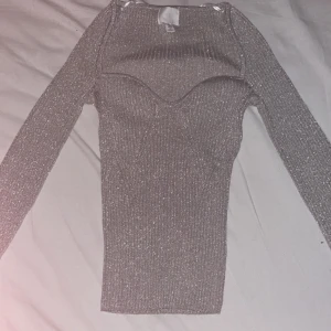 Glittrig grå topp från H&M - Säljer en superfin glittrig grå topp från H&M i storlek S. Den har en tight passform och långa ärmar, perfekt för festliga tillfällen. Toppen är ribbad och har en snygg detalj vid halsringningen. Perfekt för att glittra på festen! ✨