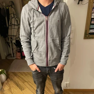 Grå hoodie från Tommy Hilfiger - Säljer en grå hoodie från Tommy Hilfiger med dragkedja. Den har en liten reva vid fickan men är annars i bra skick. 🙌Modellen är 180 och väger 68 kg🙌 